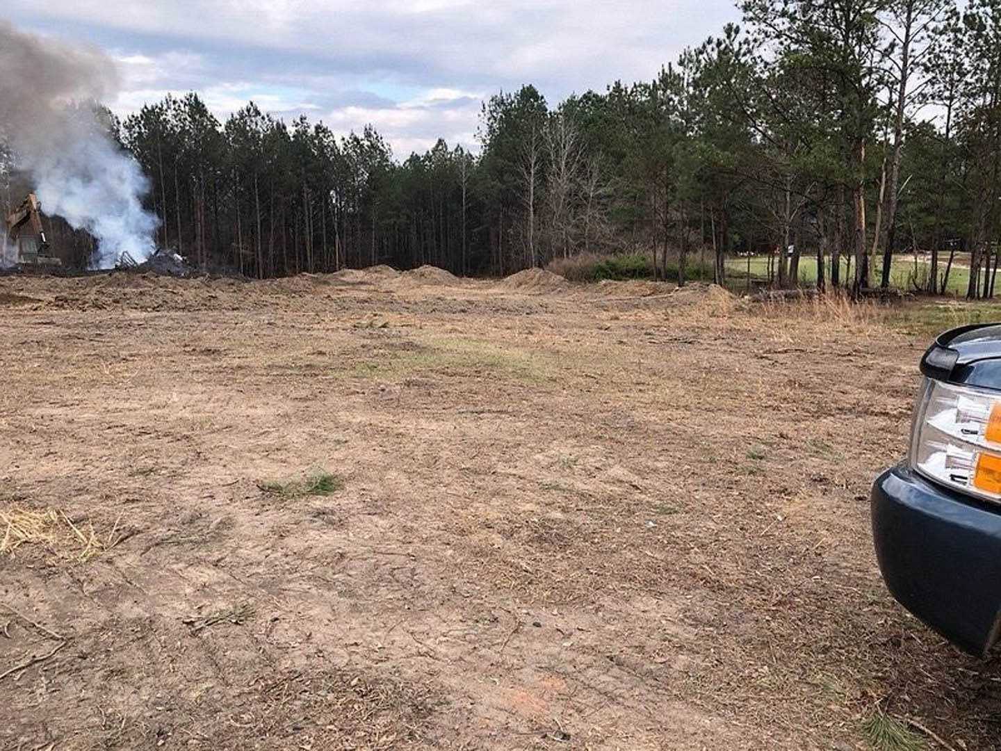 Expansive grassy field bordered by mature trees, smoke rising near a distant house, parked car partially visible in foreground
