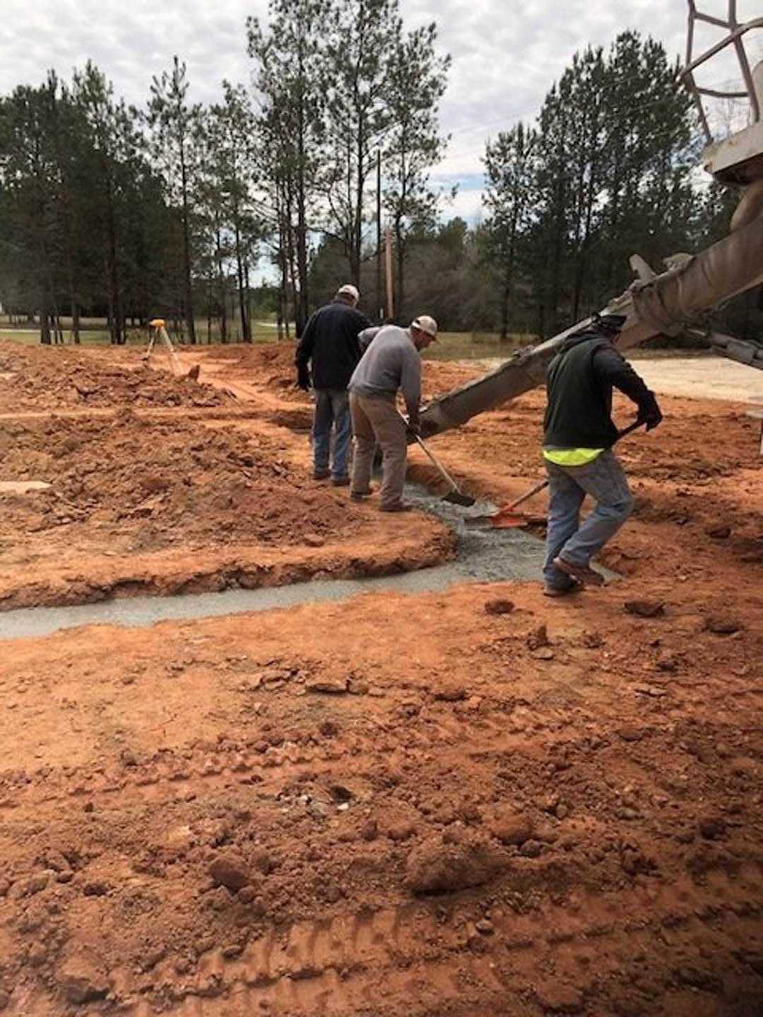 Construction workers in boots and jackets digging in muddy soil beside a partially built custom home, surrounded by trees and exposed ground under a cloudy sky