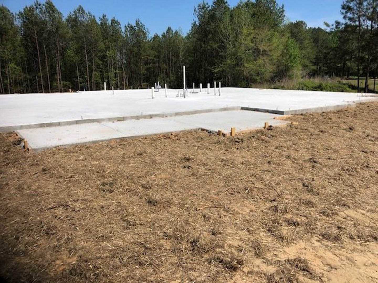 Framed custom home under construction with exposed wood beams, concrete foundation, grassy lot, and mature trees in the background