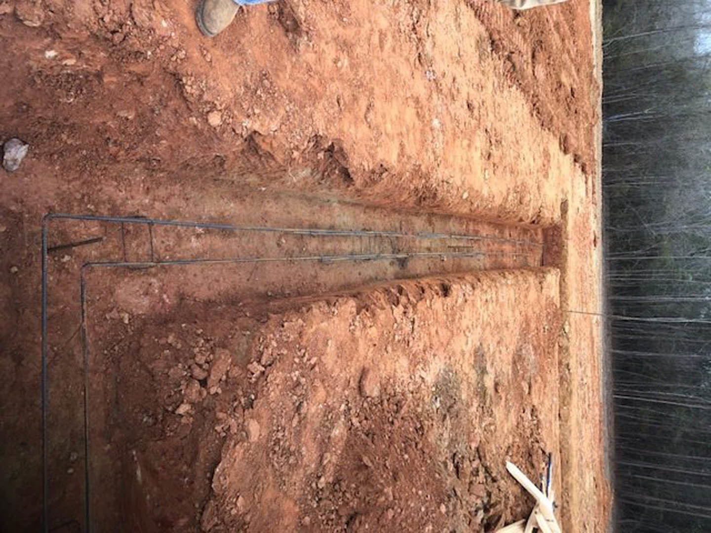 Construction site with exposed wood framing, brown dirt ground, several people standing near unfinished walls, visible wire in soil, close-up of window and white construction