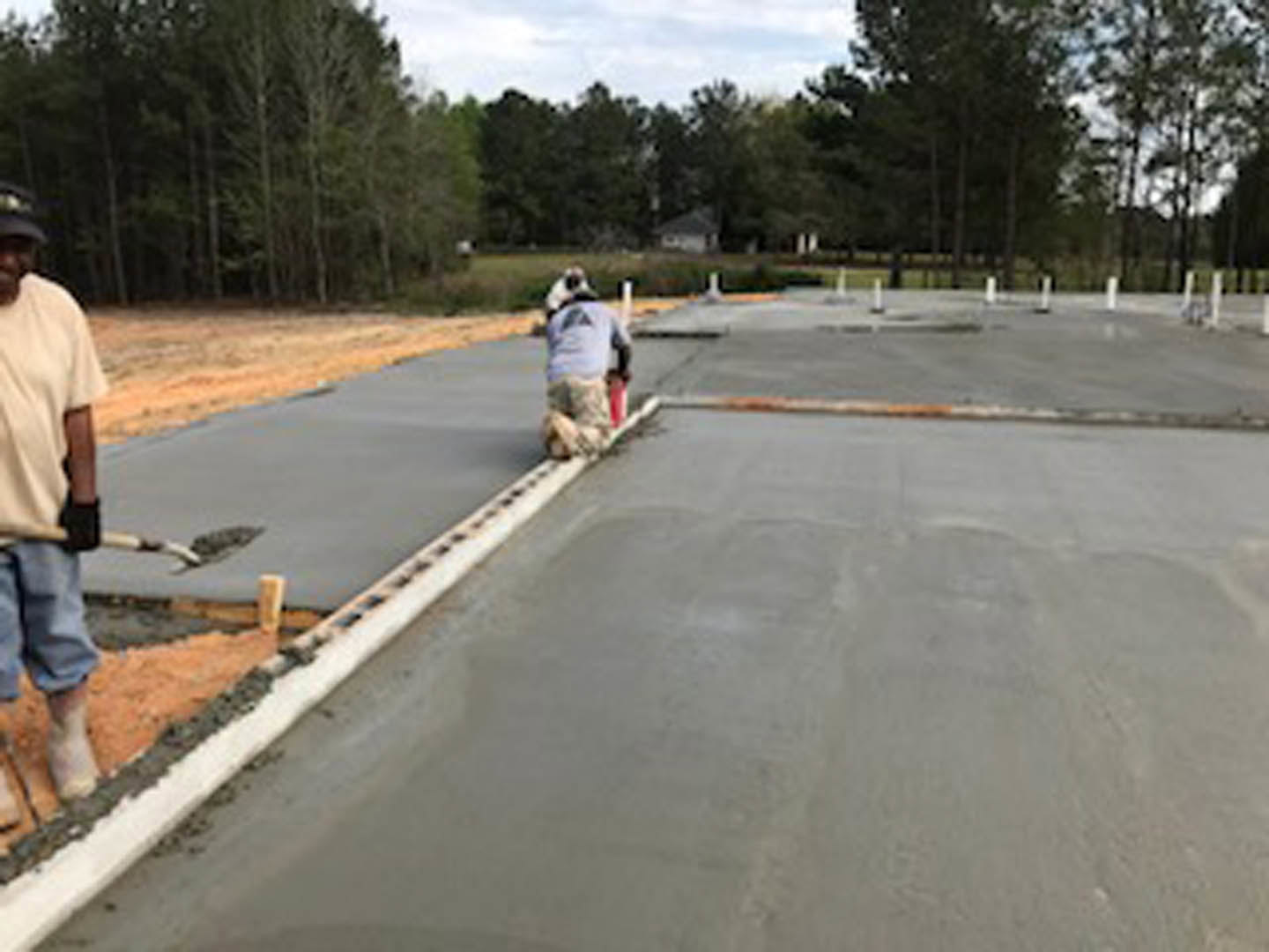 Modern home exterior with light stone facade, large windows, and landscaped front yard bordered by mature trees; paved driveway and sidewalk visible in foreground.