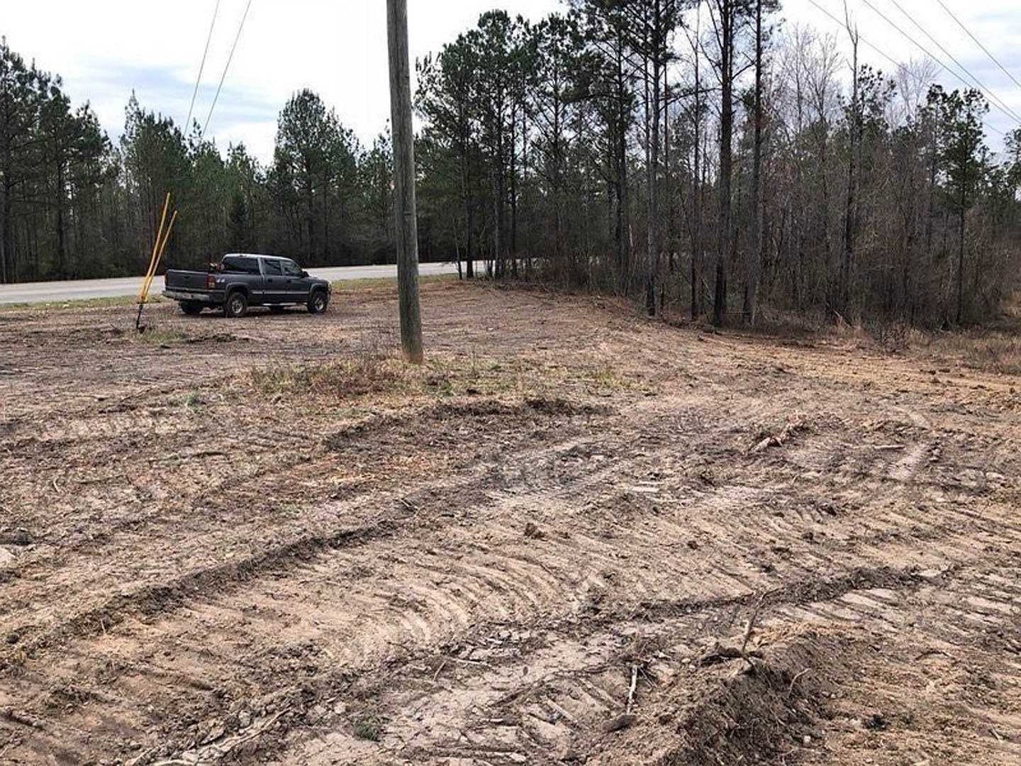 Black pickup truck parked on a dirt lot beside grassy area, tire tracks visible in soil, trees and blue sky in background