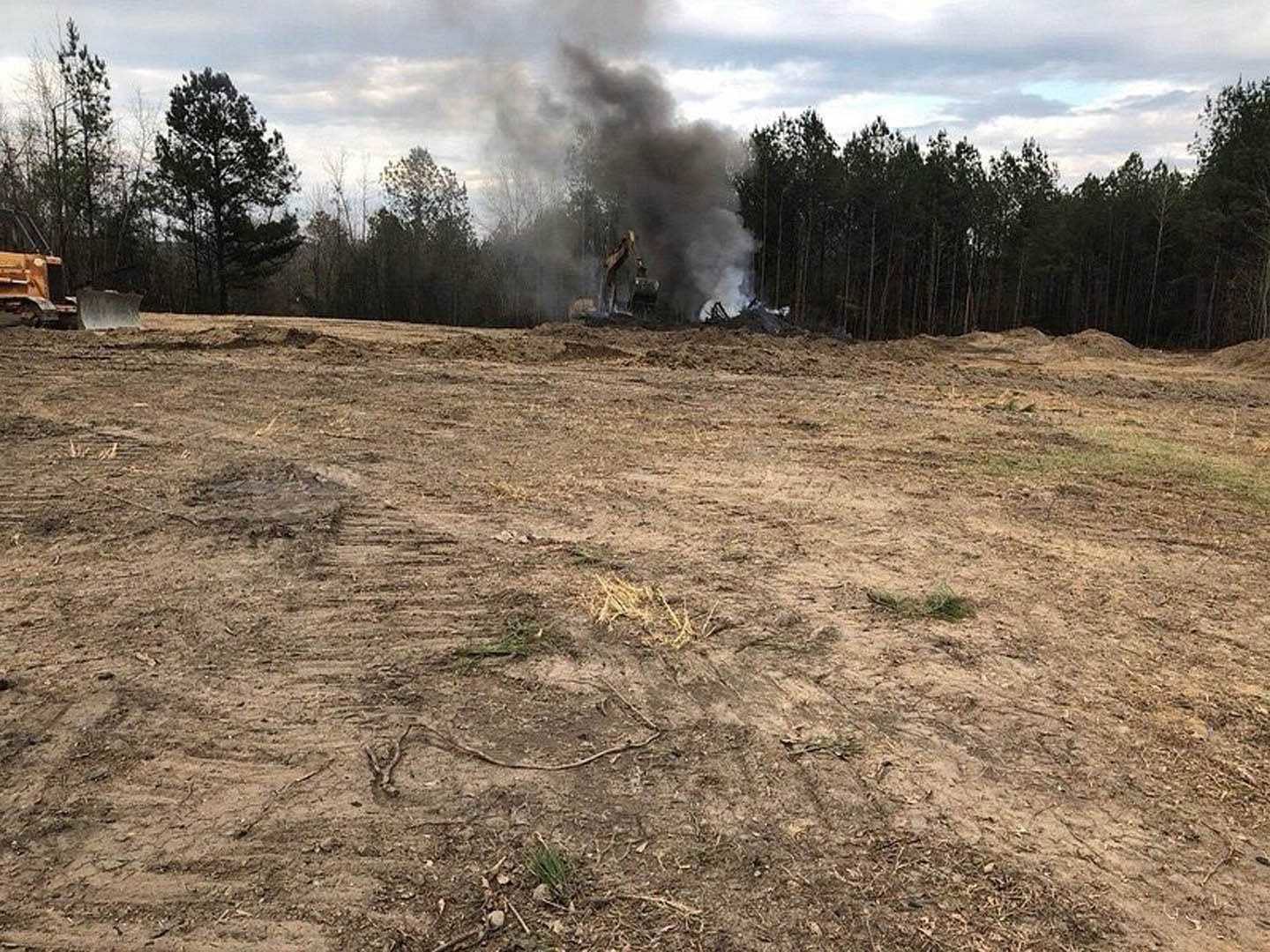 Dirt field with tire tracks, burning truck emitting thick black smoke, grassy edges and scattered trees in background