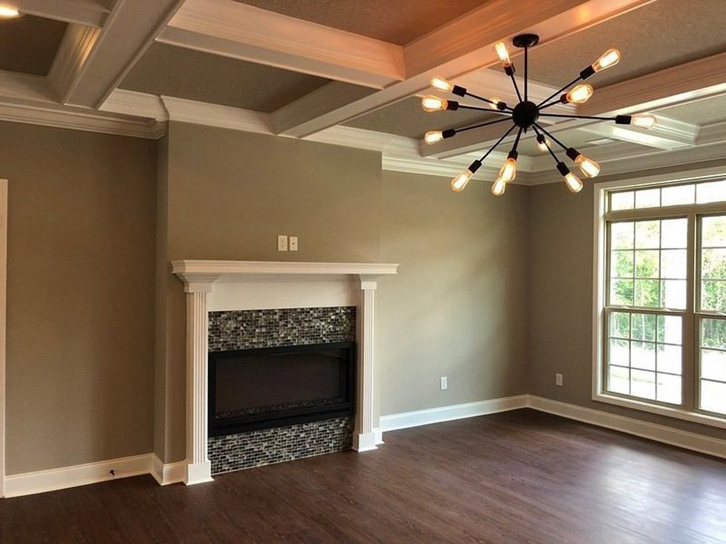 Living room with multi-pane window, brick fireplace, wood flooring, and ornate chandelier hanging from a white ceiling with crown molding.