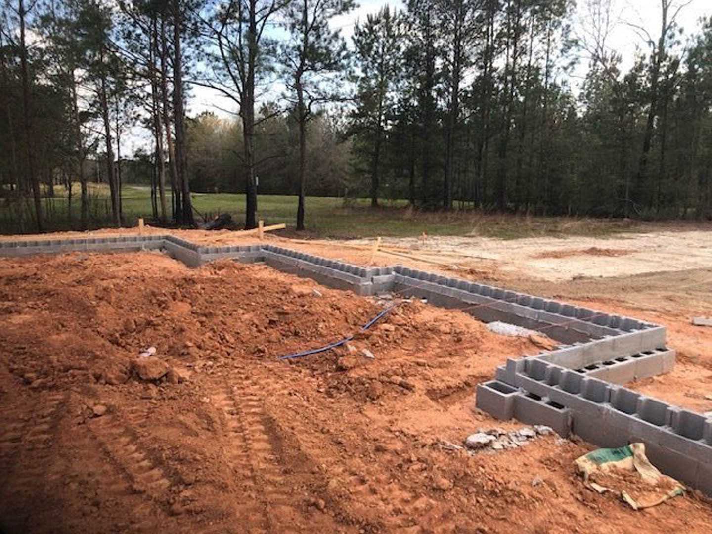 Concrete foundation forms set in packed dirt, tire tracks visible in soil, scattered construction debris, trees lining background