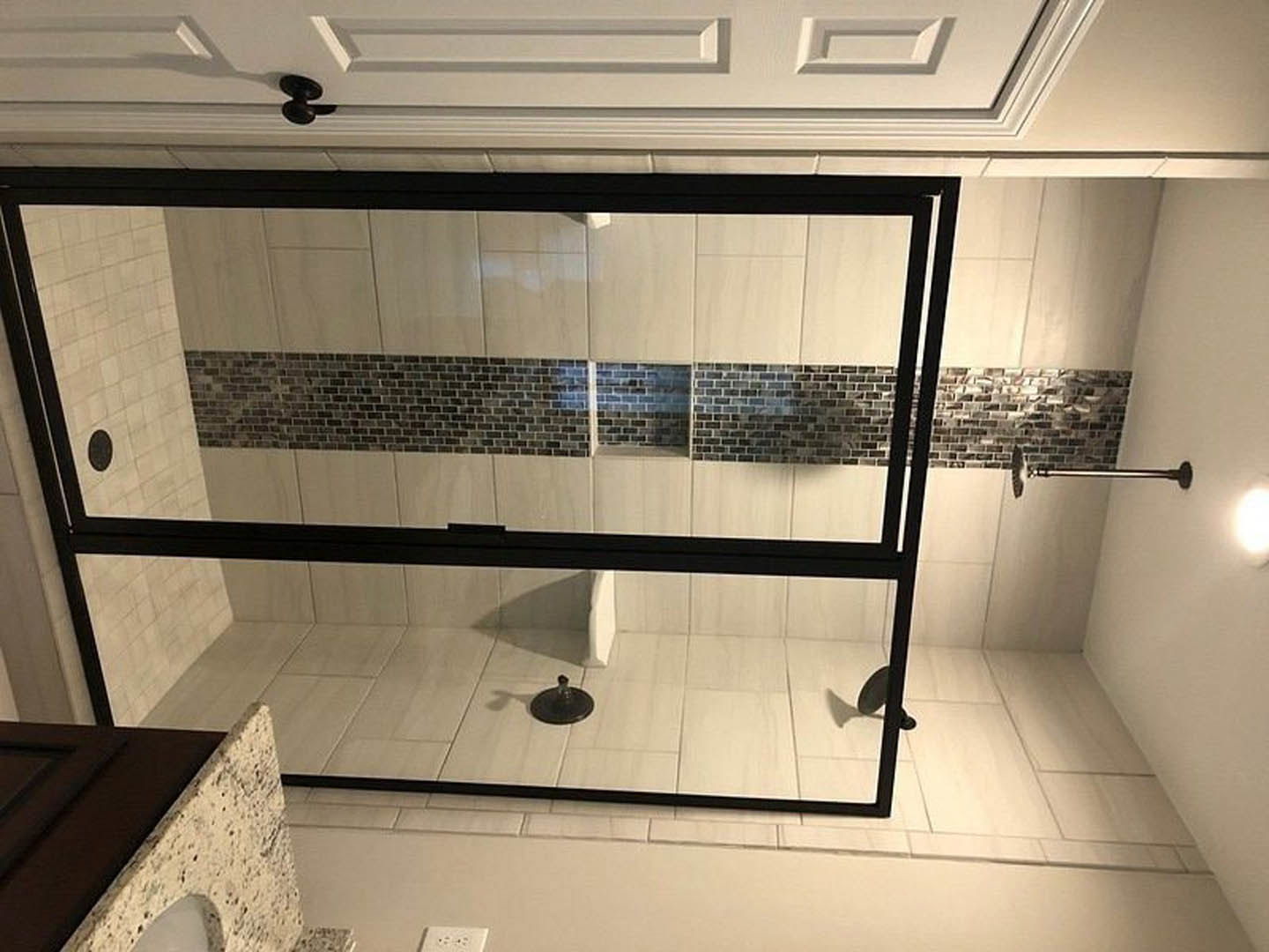 Walk-in shower with clear glass doors, white tile walls, black metal fixtures, adjacent white vanity with sink, black hardware, and a white door with black handle