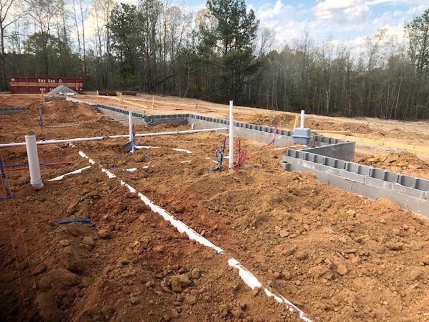 Partially built home foundation surrounded by dirt, white pipes, and scattered trees under a blue sky with clouds