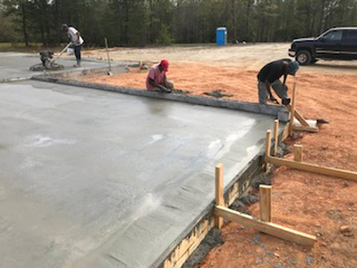 Crew pouring and leveling concrete slab foundation beside parked black SUV, surrounded by trees and construction materials