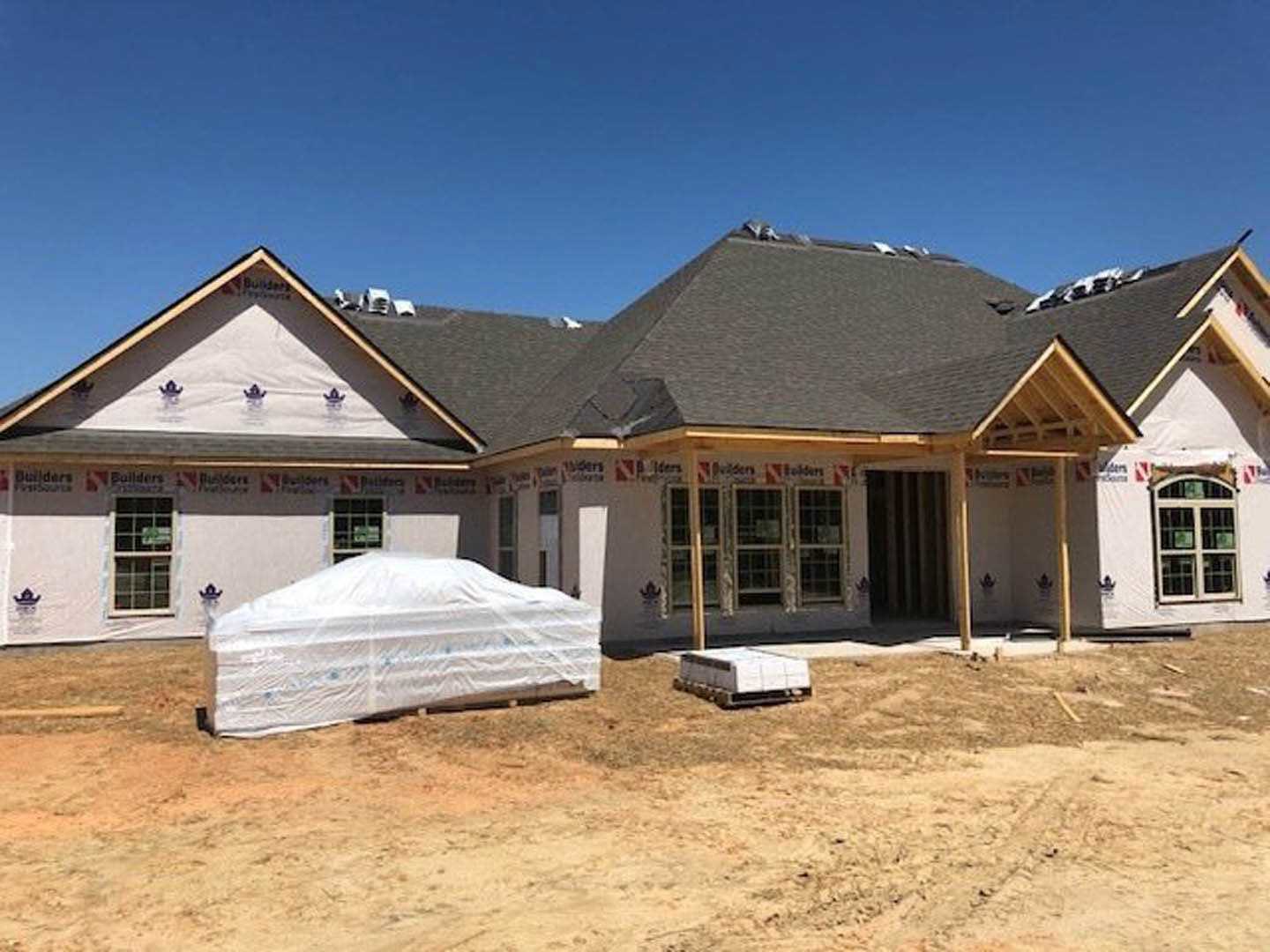 Partially built house with exposed wooden framing, large roof, and blue tarp covering construction materials; white car parked nearby with protective cover; close-up of window