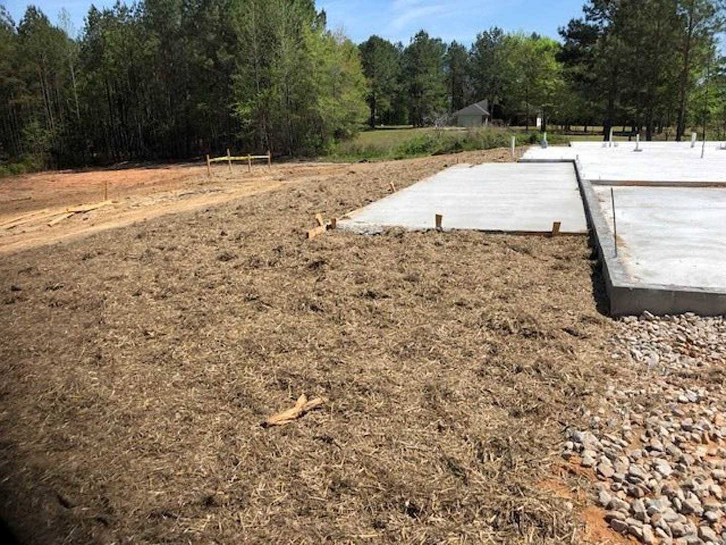 Concrete slab foundation with exposed metal rods set in a grassy field, surrounded by soil and scattered trees under an open sky