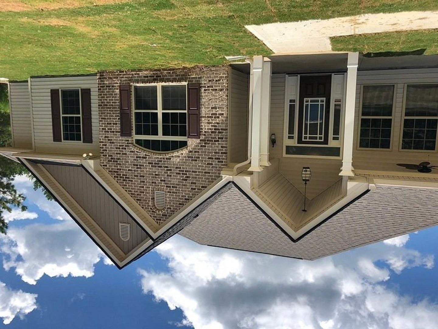 Two-story home with gray siding, black front door with white trim, large white-framed windows, shingle roof, manicured green lawn, small tree, and blue sky with scattered clouds