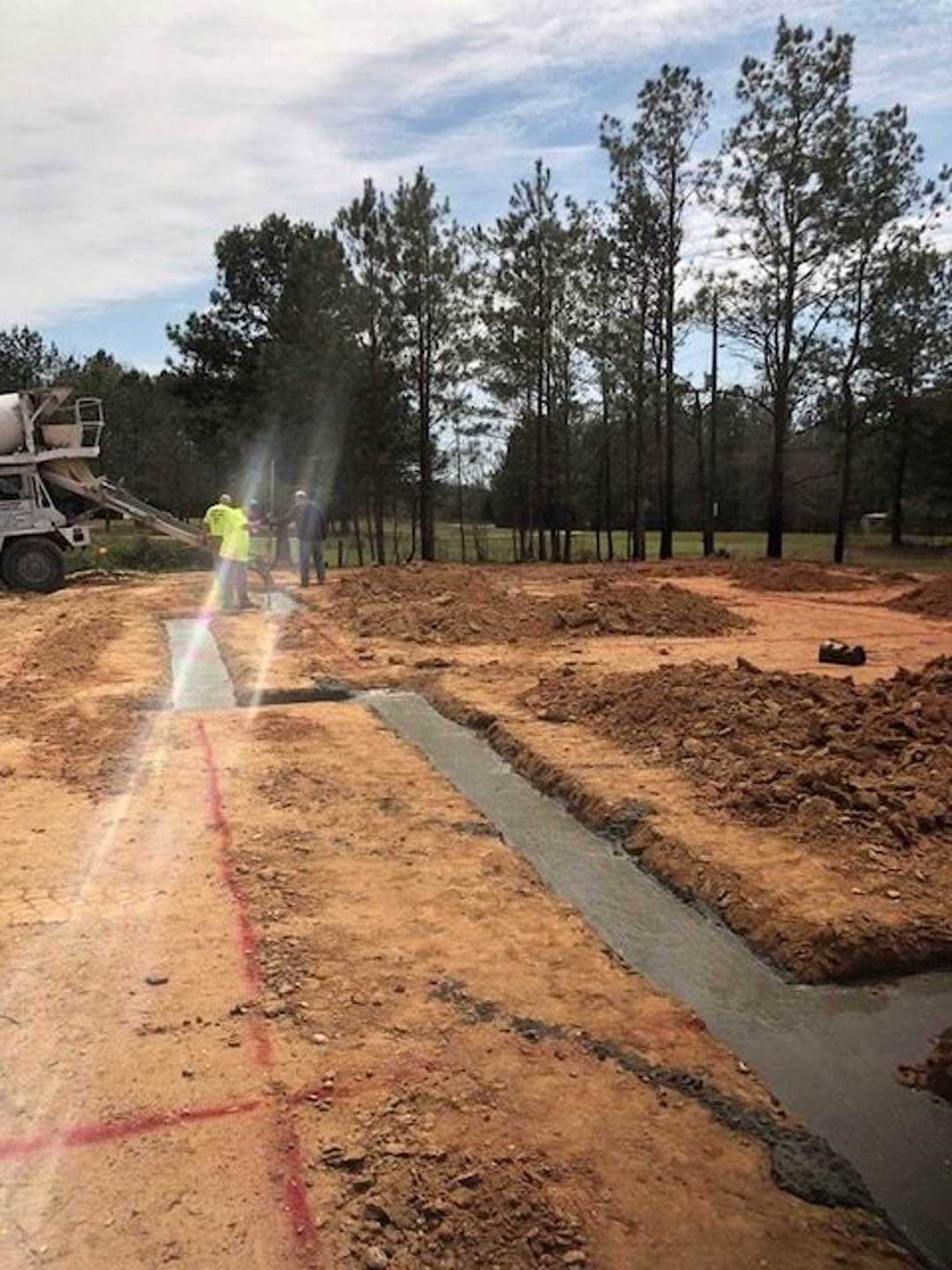 Construction site with two trucks, one featuring a cement mixer, dirt ground, scattered soil, trees and plants in the background, cloudy sky overhead, blurry figure in a yellow
