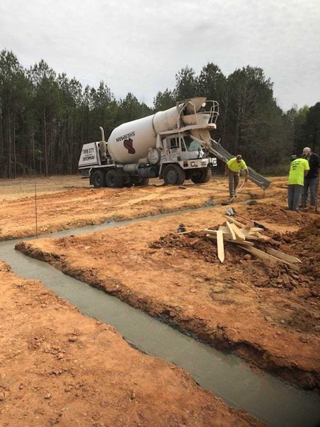 Construction site with white cement mixer truck parked on dirt, several workers in yellow shirts, grassy area and trees in background