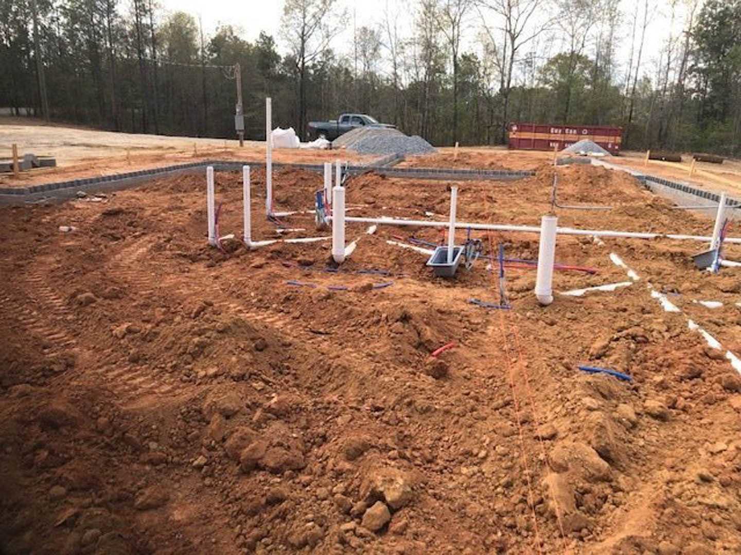 White PVC pipes laid across soil at a residential construction site, with a pickup truck and trees in the background