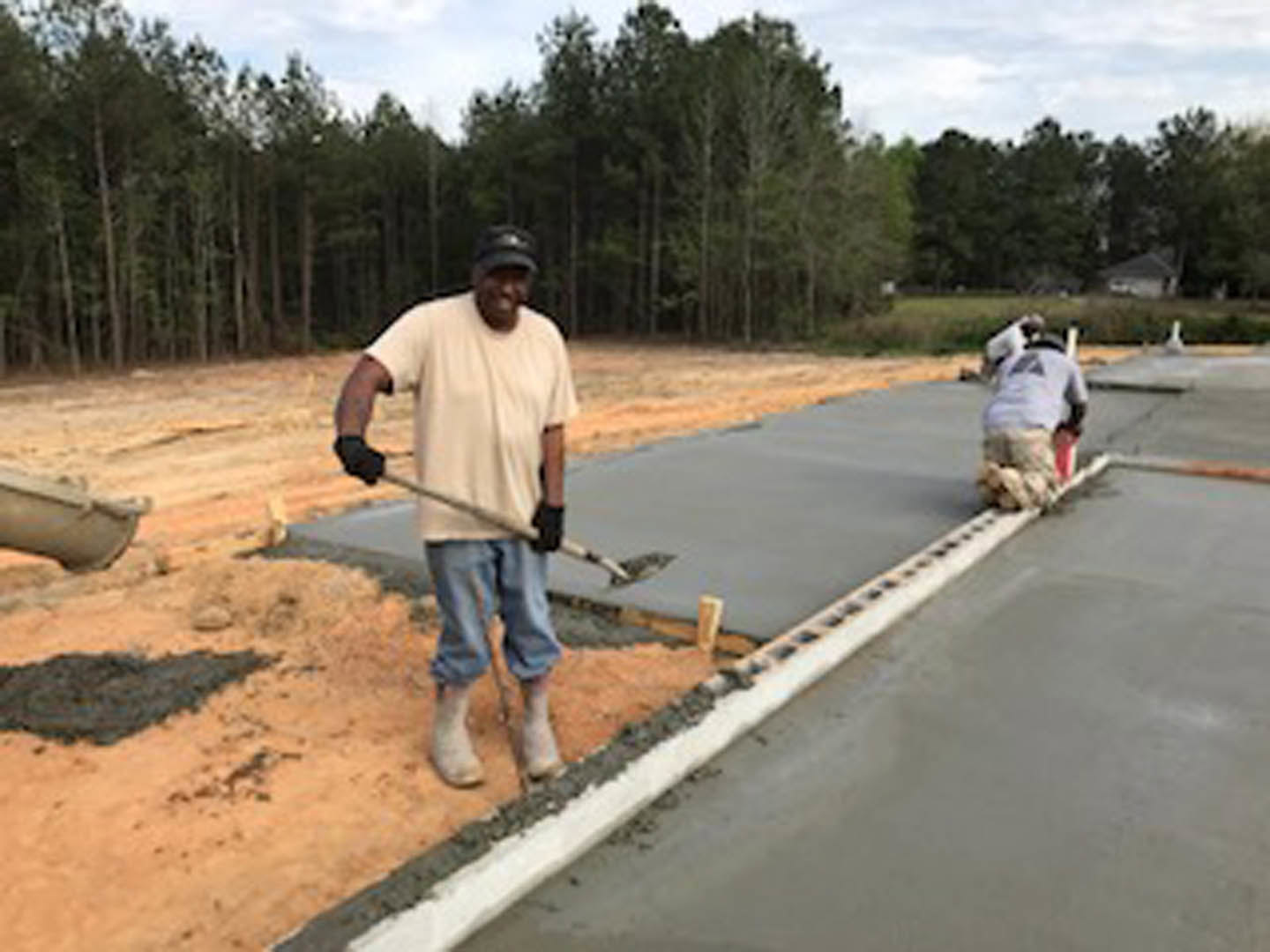 Man in work gloves and boots stands on dirt foundation outside custom home, holding shovel near construction materials; trees and sky visible in background.