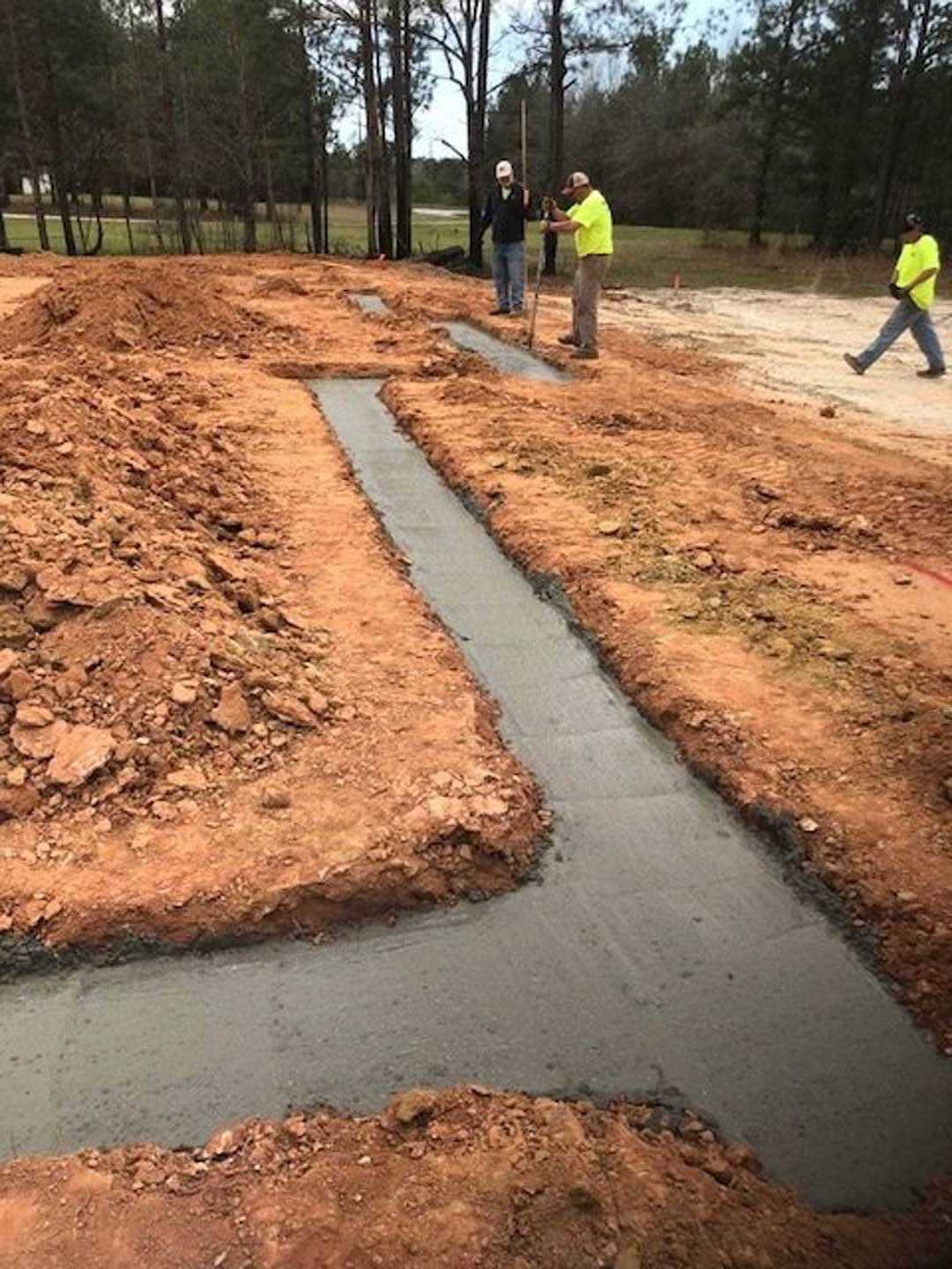 Several men standing on bare soil outside, surrounded by grass, trees, and plants under a clear sky