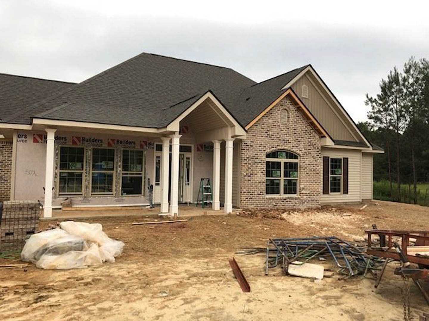 Two-story house under construction with exposed pillars, metal scaffolding stacked in front, multi-pane windows, plastic bags on the ground, and trees in the background