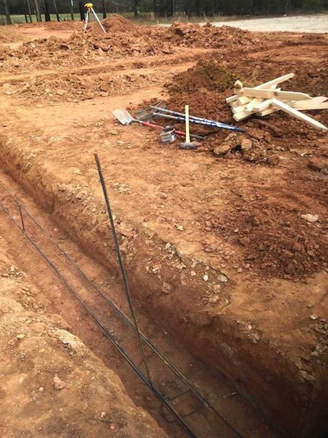 Construction site with scattered metal rods and wood pieces on bare soil, surrounded by trees and muddy ground.