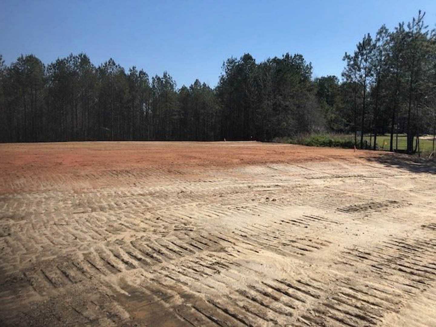 Brown dirt field with tire tracks, bordered by dense green trees under a clear sky