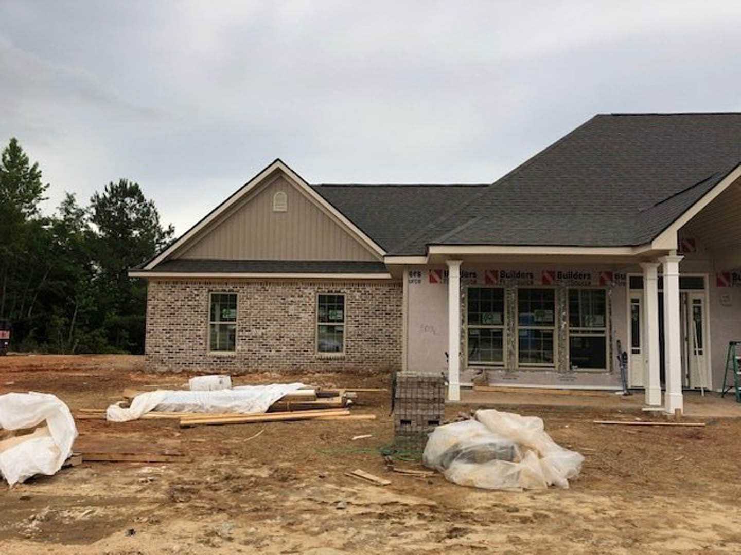 Wood-framed house under construction with scattered debris on bare ground, partially installed multi-pane windows, plastic bag with dark object near foundation, trees in background