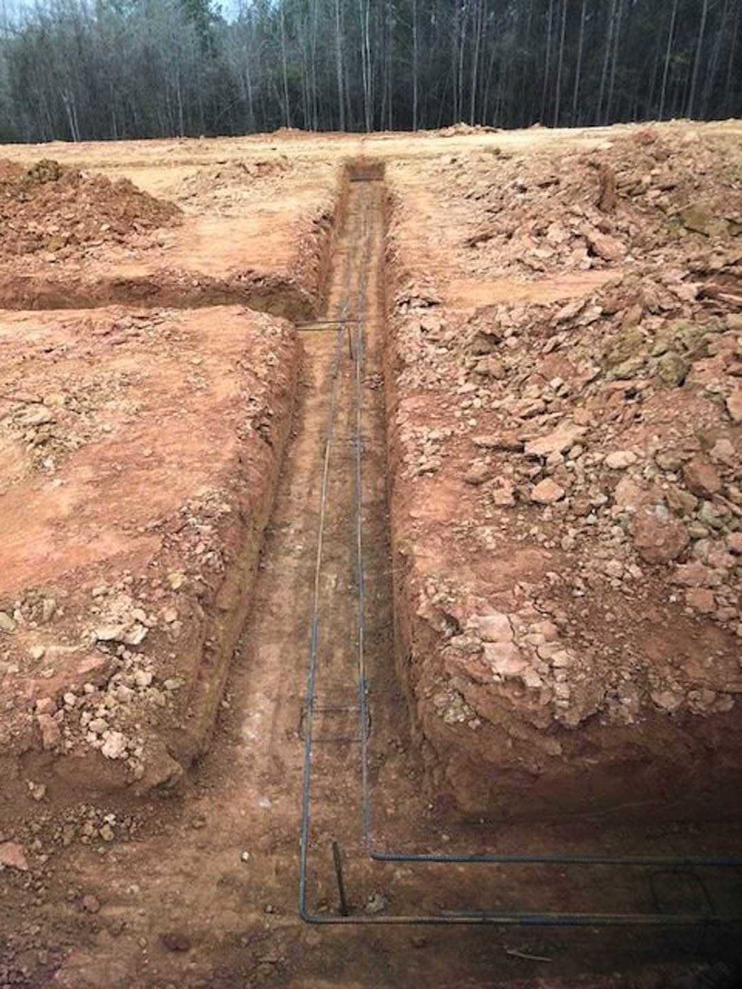Freshly dug trench in a dirt field with exposed soil, bordered by scattered trees and vegetation.