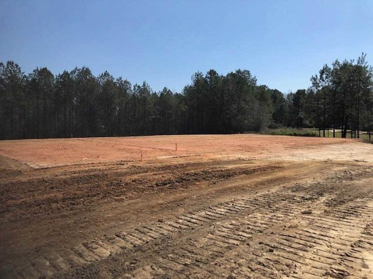 Dirt field with tire tracks bordered by a group of trees under a clear blue sky