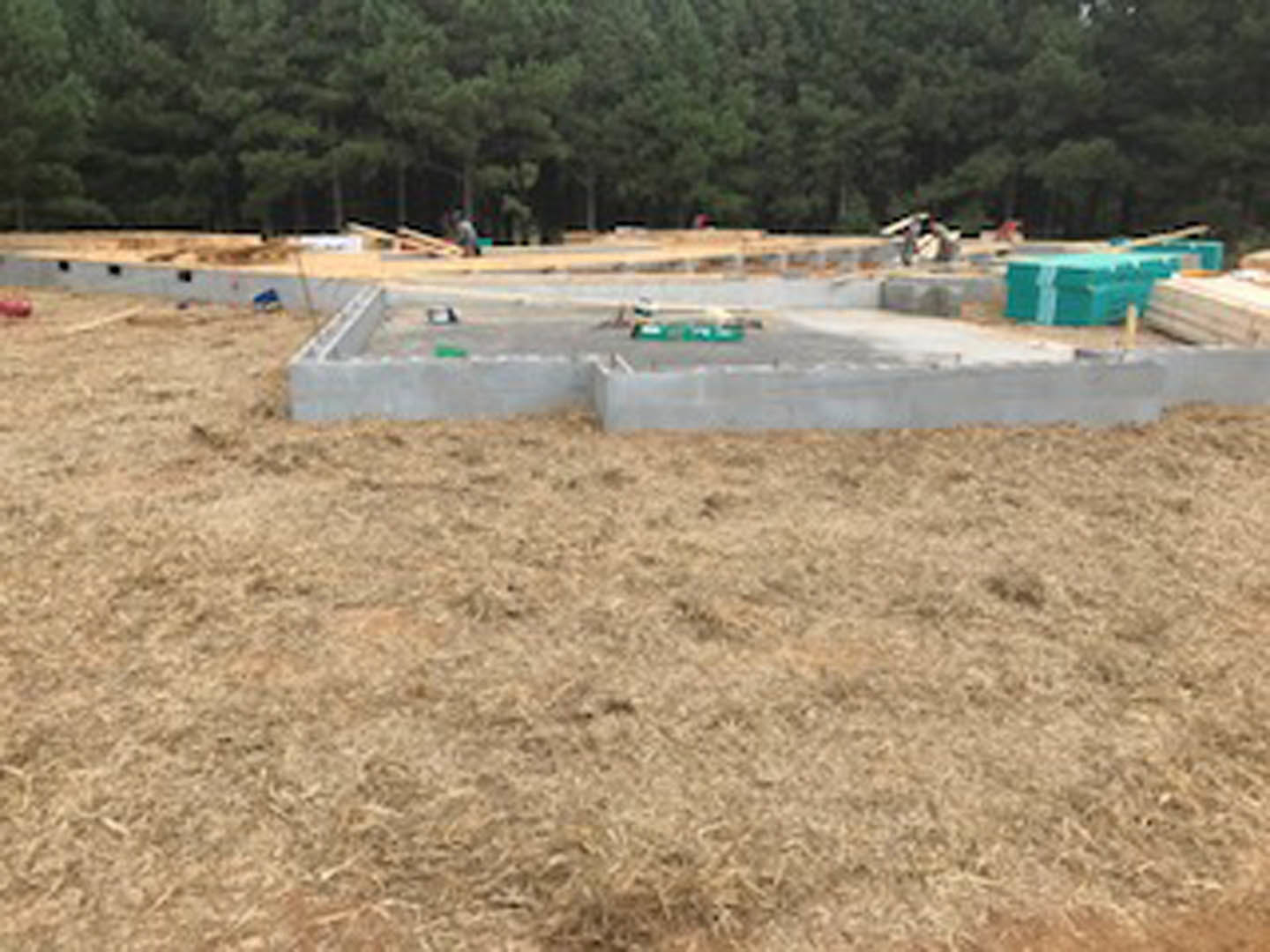 Concrete foundation surrounded by grass and soil, several people standing on the building site, trees in the background