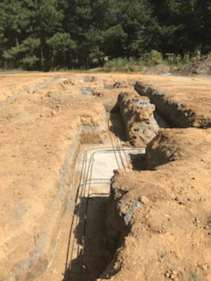 Excavated dirt pit surrounded by metal rods and fencing, with trees and dense greenery in the background