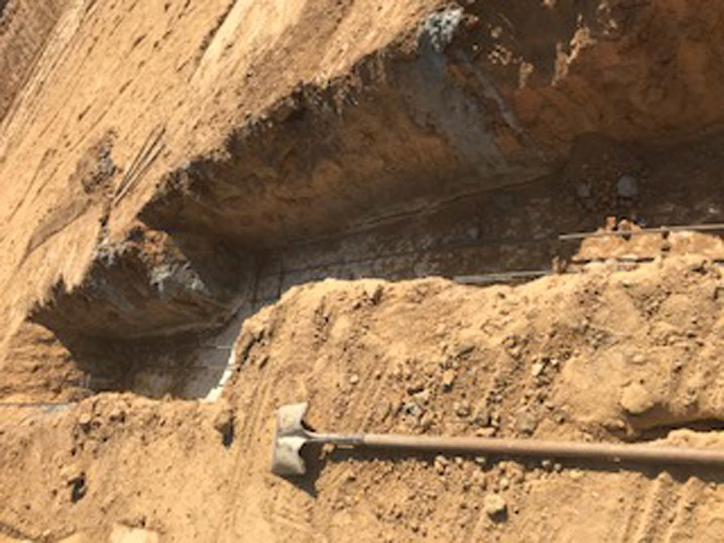 Shovel resting on bare dirt beside rough rock wall and exposed pipe, outdoor ground with scattered stones