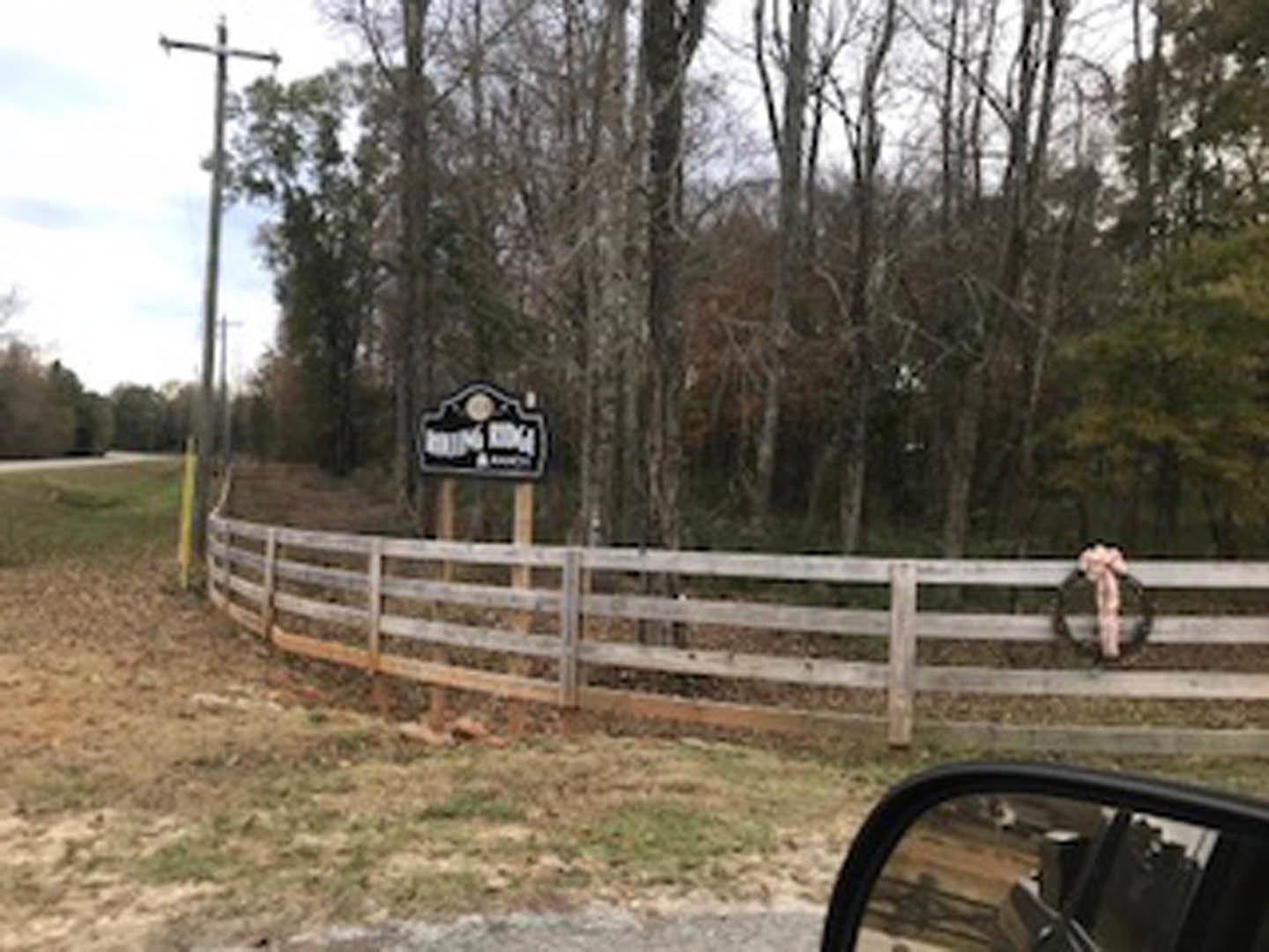 Split rail fence with attached sign, grassy yard, mature trees, and blue sky in background; partial view of car side mirror and blurred leg in foreground