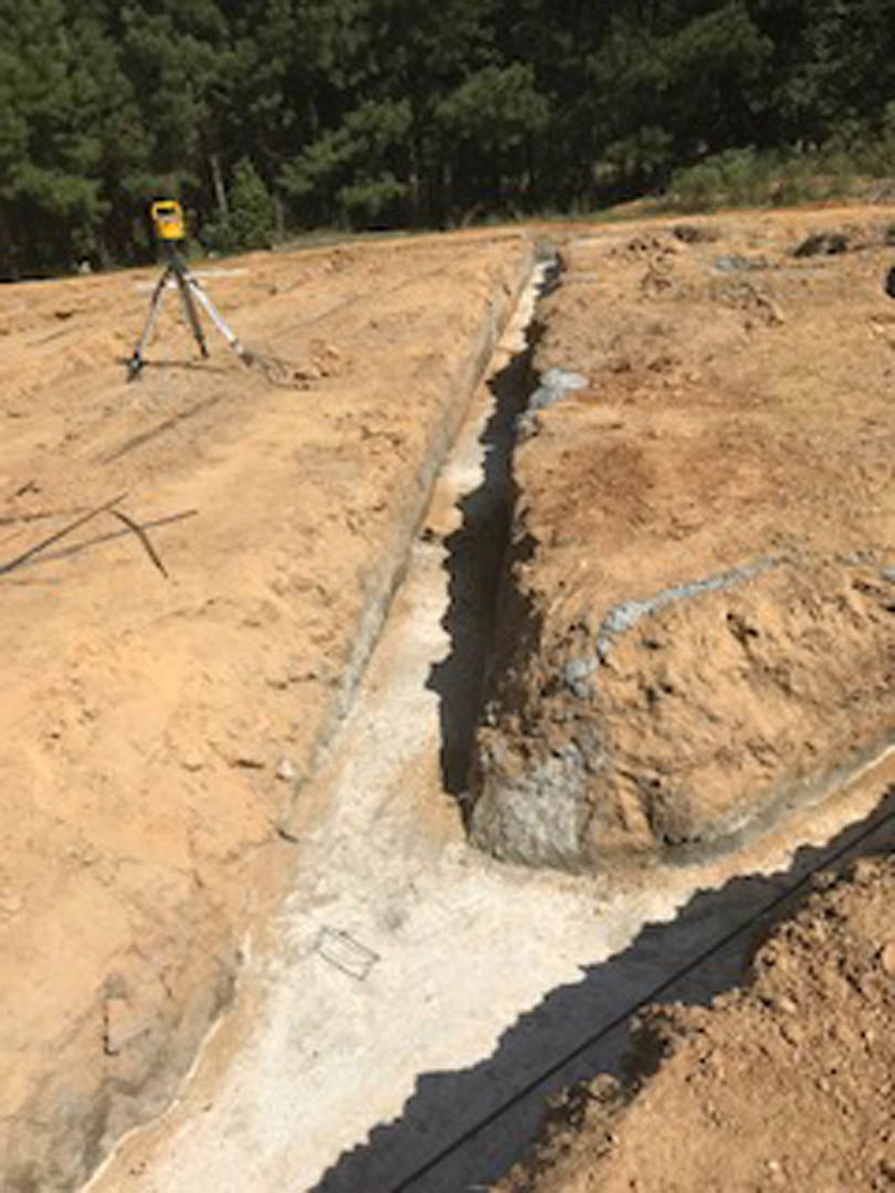 Land survey equipment mounted on a tripod positioned on a dirt and rocky ground near trees and soil, with a man standing in the background