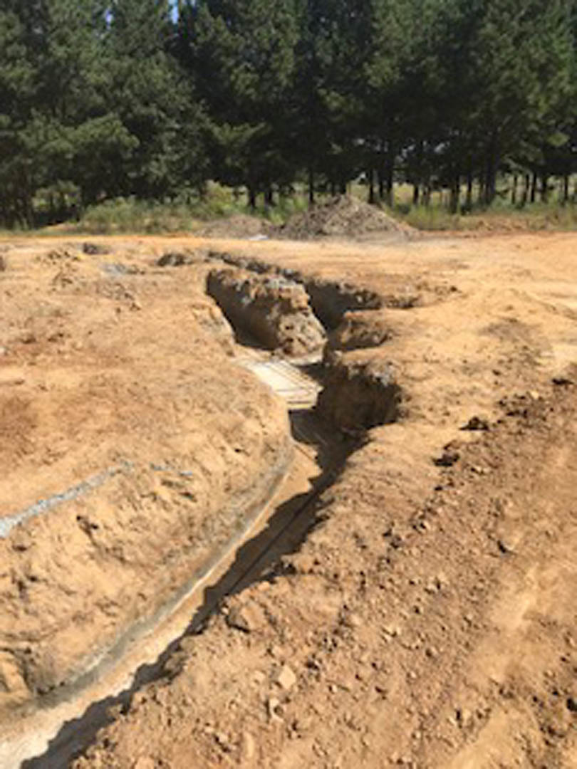 Excavated construction site with exposed soil, piles of dirt and grass, surrounded by trees and open landscape