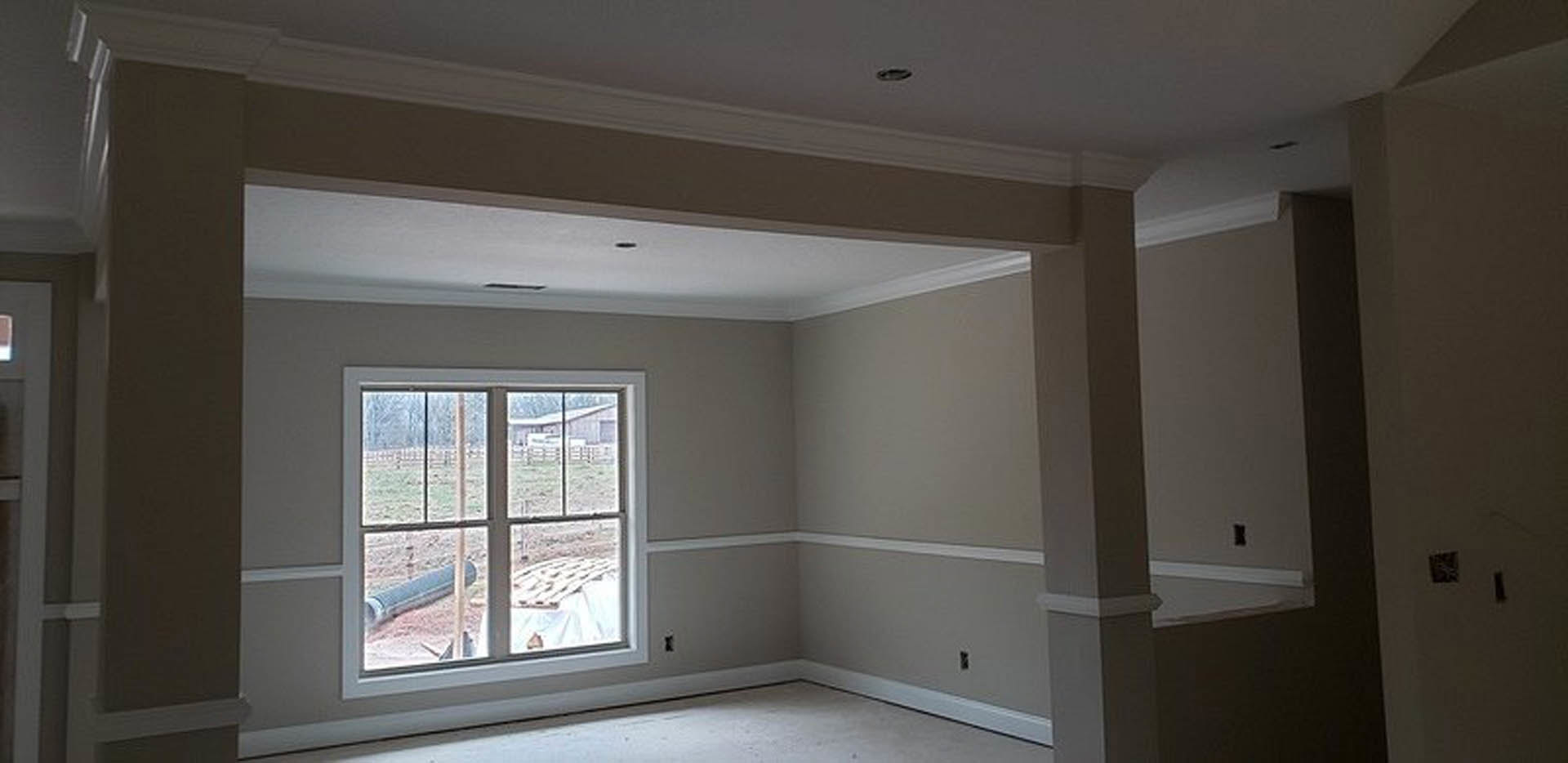 White plaster walls with decorative molding, large window overlooking farmland, ceiling with recessed lighting, square thermostat mounted on wall, exposed metal pipe near window