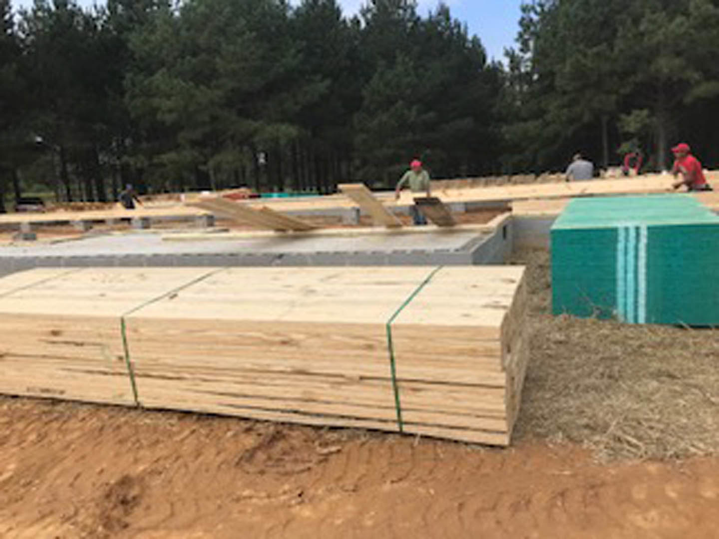 Workers assembling wooden foundation beams on a residential construction site with soil, trees, and blue sky in the background