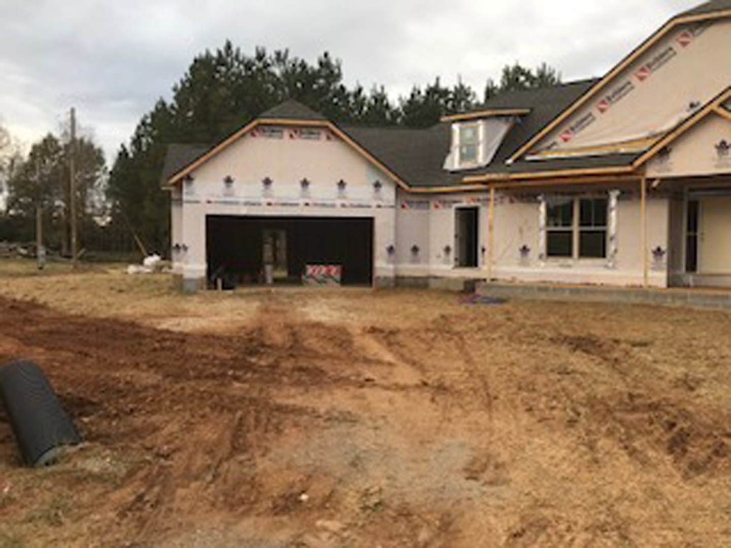 Two-story house under construction with exposed framing, surrounded by dirt lot and mature trees, cloudy sky overhead
