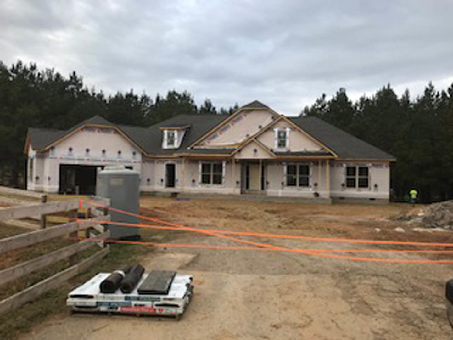 Two-story house with black roof, white siding, and wooden privacy fence; driveway with parked car, green lawn, mature trees, and partly cloudy sky in background