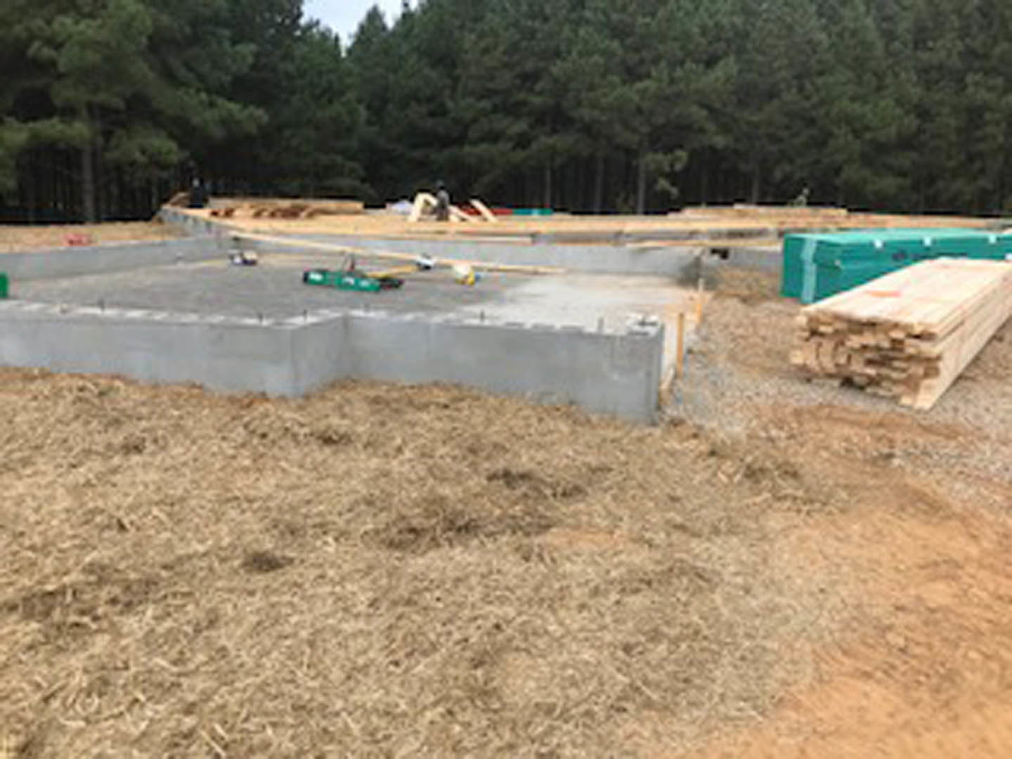 Construction site with stacked wood planks, concrete foundation blocks, grassy field, and trees in the background