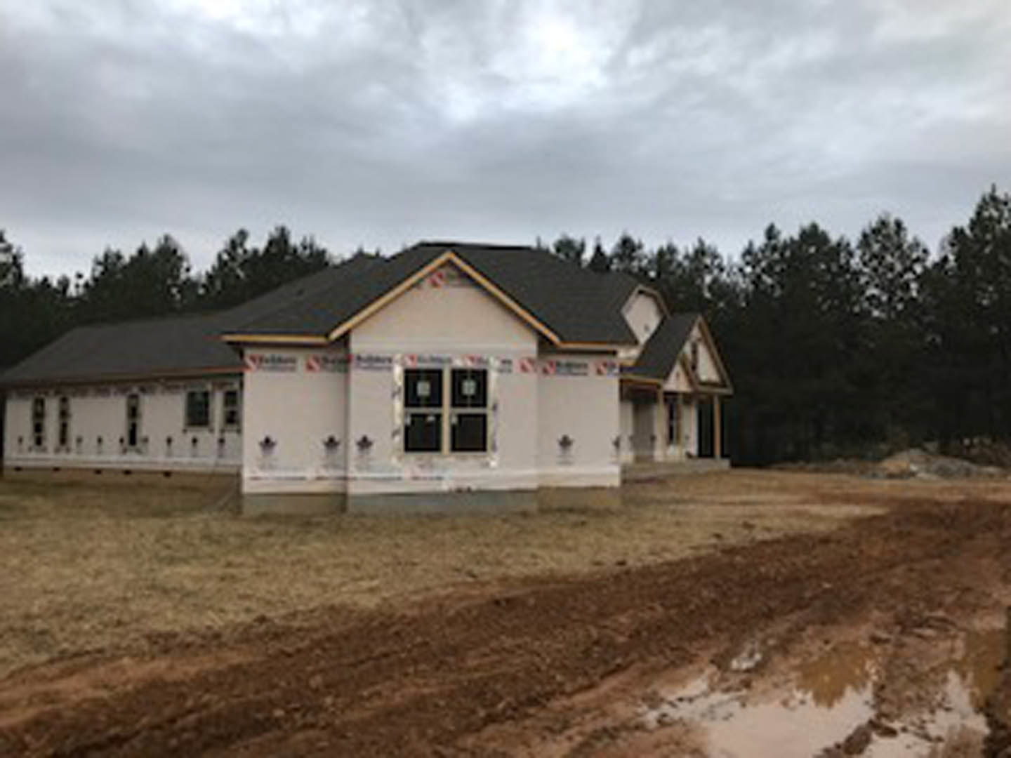 Partially built white house with exposed framing, surrounded by muddy ground and mature trees under a cloudy sky