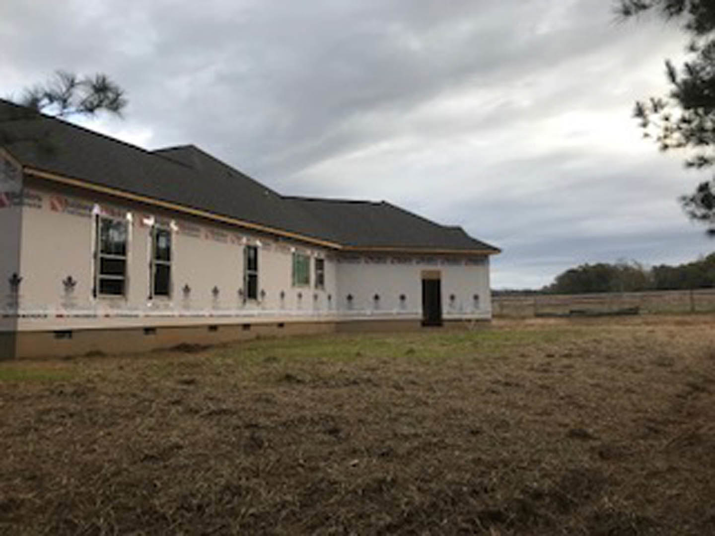 Modern cottage with black roof and white siding set on a grassy field, surrounded by trees under a cloudy sky