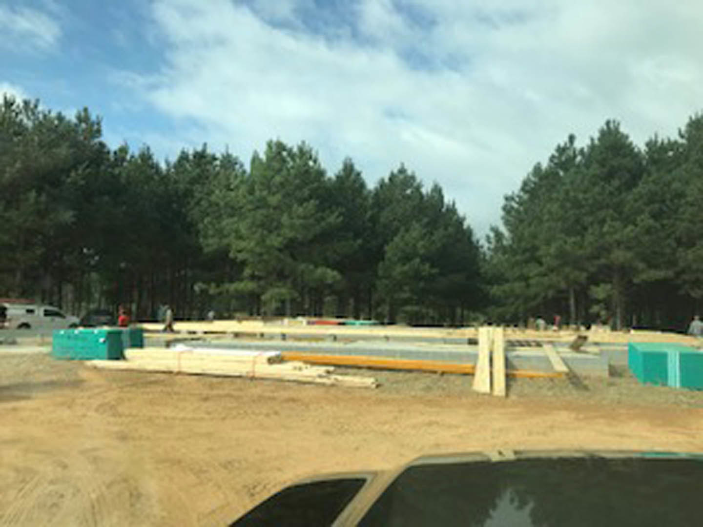 Framed custom home under construction with exposed wood beams, gravel ground, and tall trees lining the background