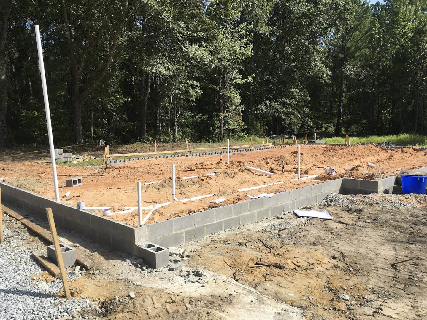 Concrete house foundation surrounded by gravel, wooden planks, and construction materials; trees and soil in the background; blue bucket and white signpost visible.