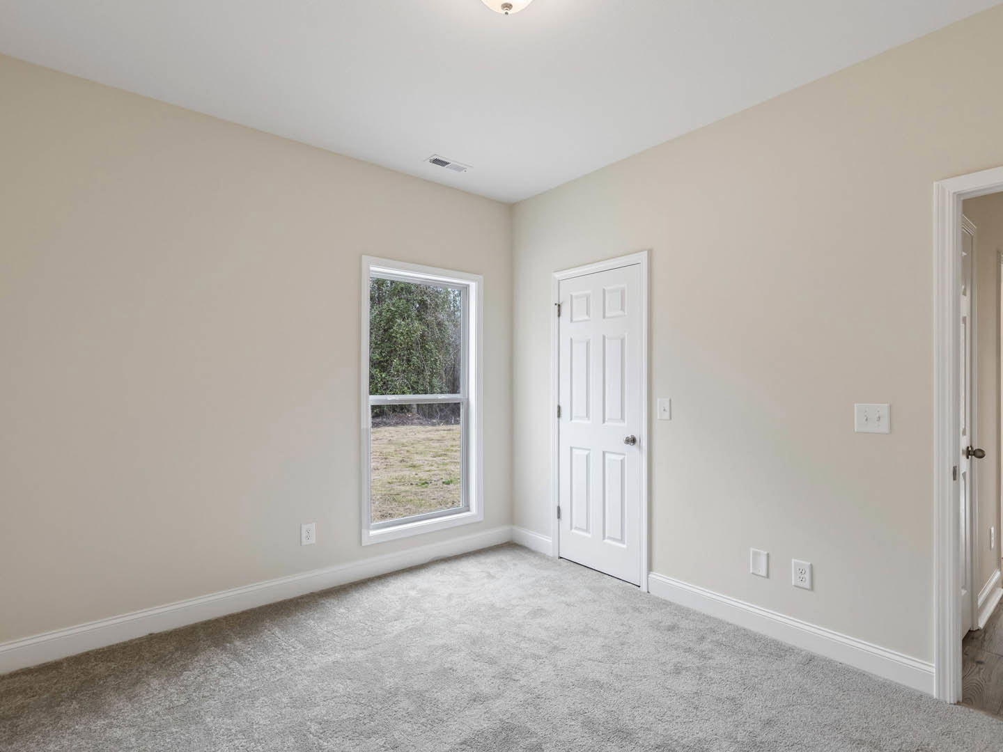 Carpeted bedroom with white door featuring silver knob, large window overlooking grassy field and trees, light-colored walls with simple molding