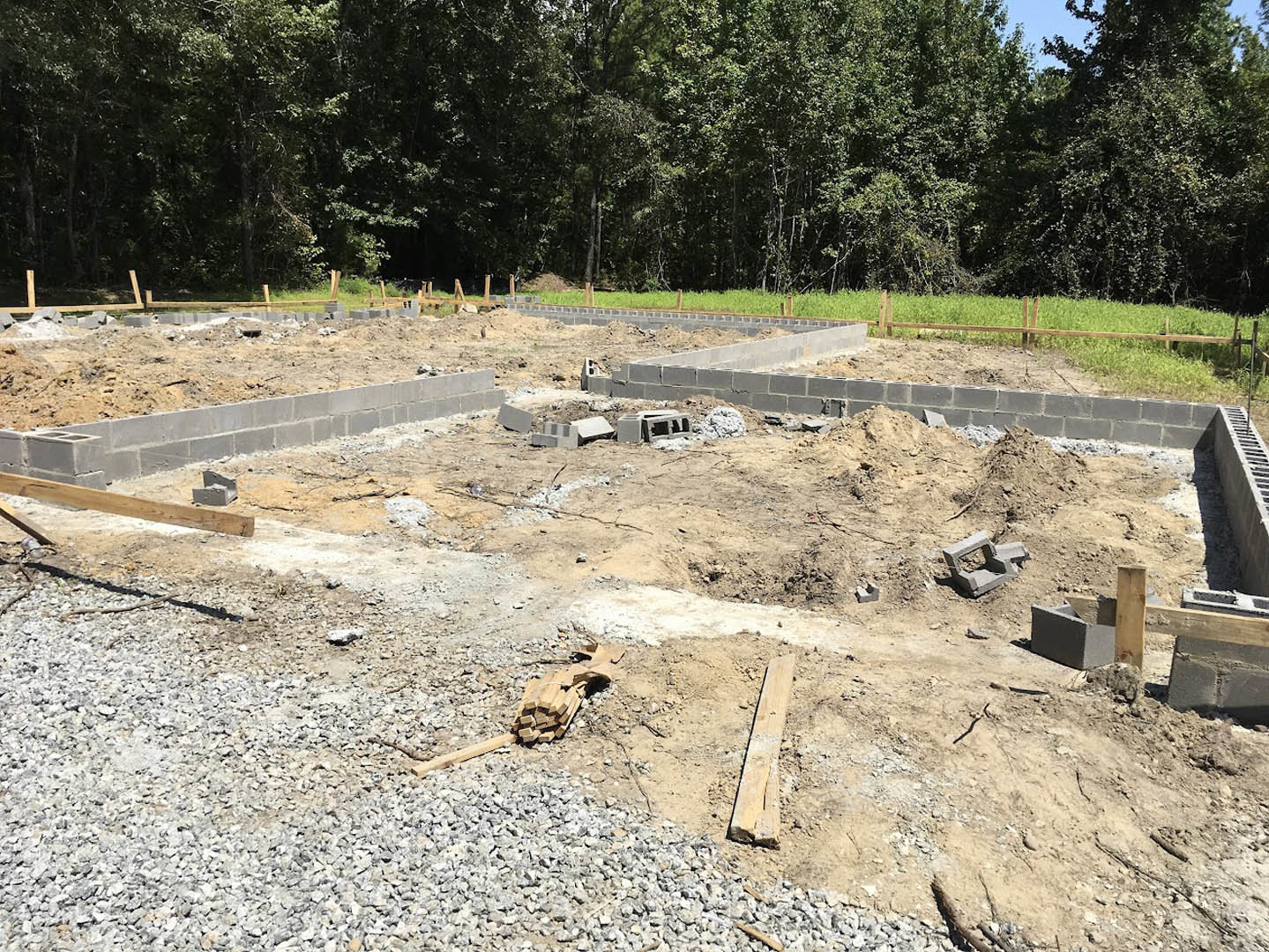 Bricks and dirt piled on a residential construction site, wooden planks and concrete blocks scattered on bare soil, surrounded by a metal fence and green trees in the background