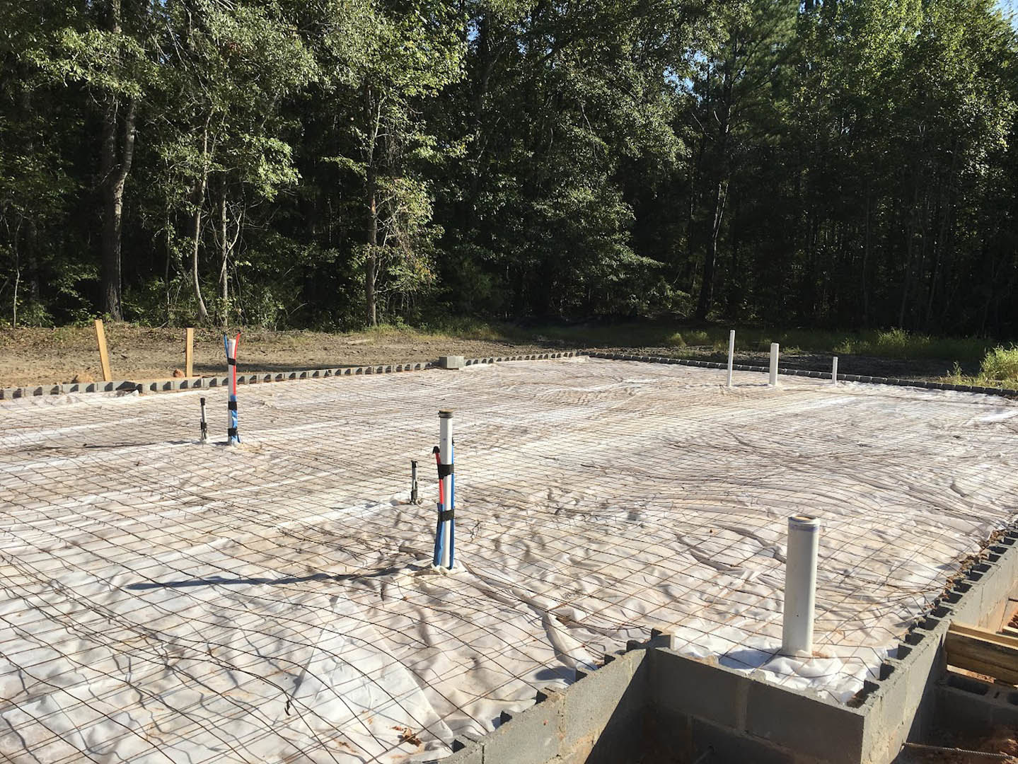 Concrete foundation wrapped in white plastic sheeting, wire mesh surrounding a vertical pole, trees and bushes in the background, white post with black stripe nearby.