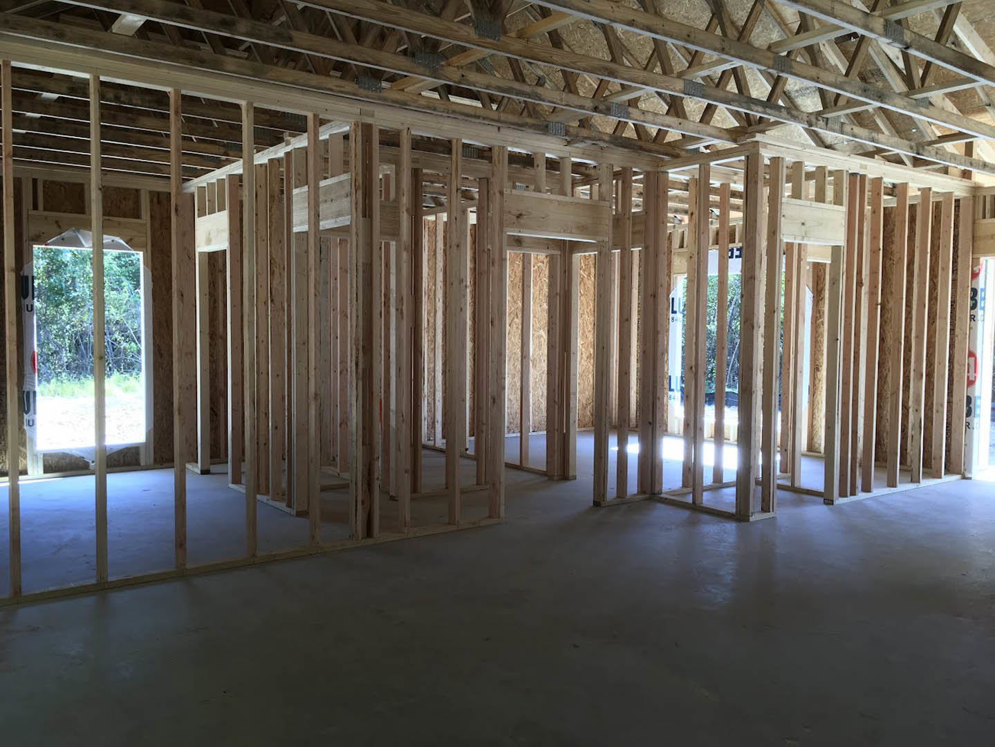Exposed wooden ceiling beams and columns over a concrete floor, large window framing tree views, unfinished interior construction