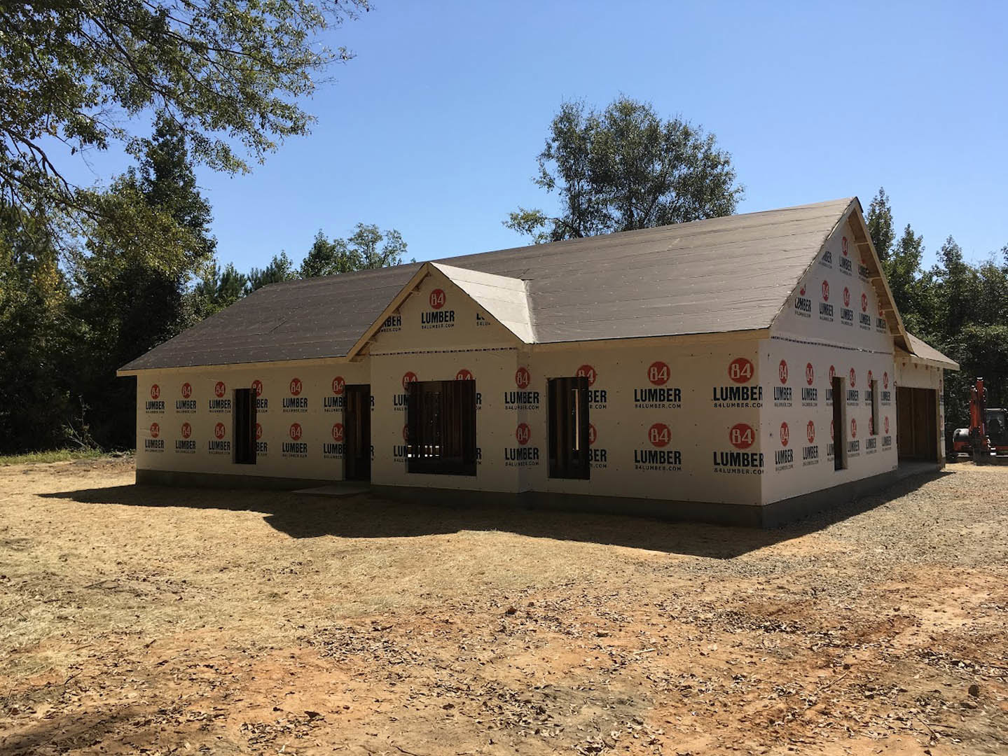 Framed house under construction with exposed plywood, open window spaces, and surrounding trees in the background