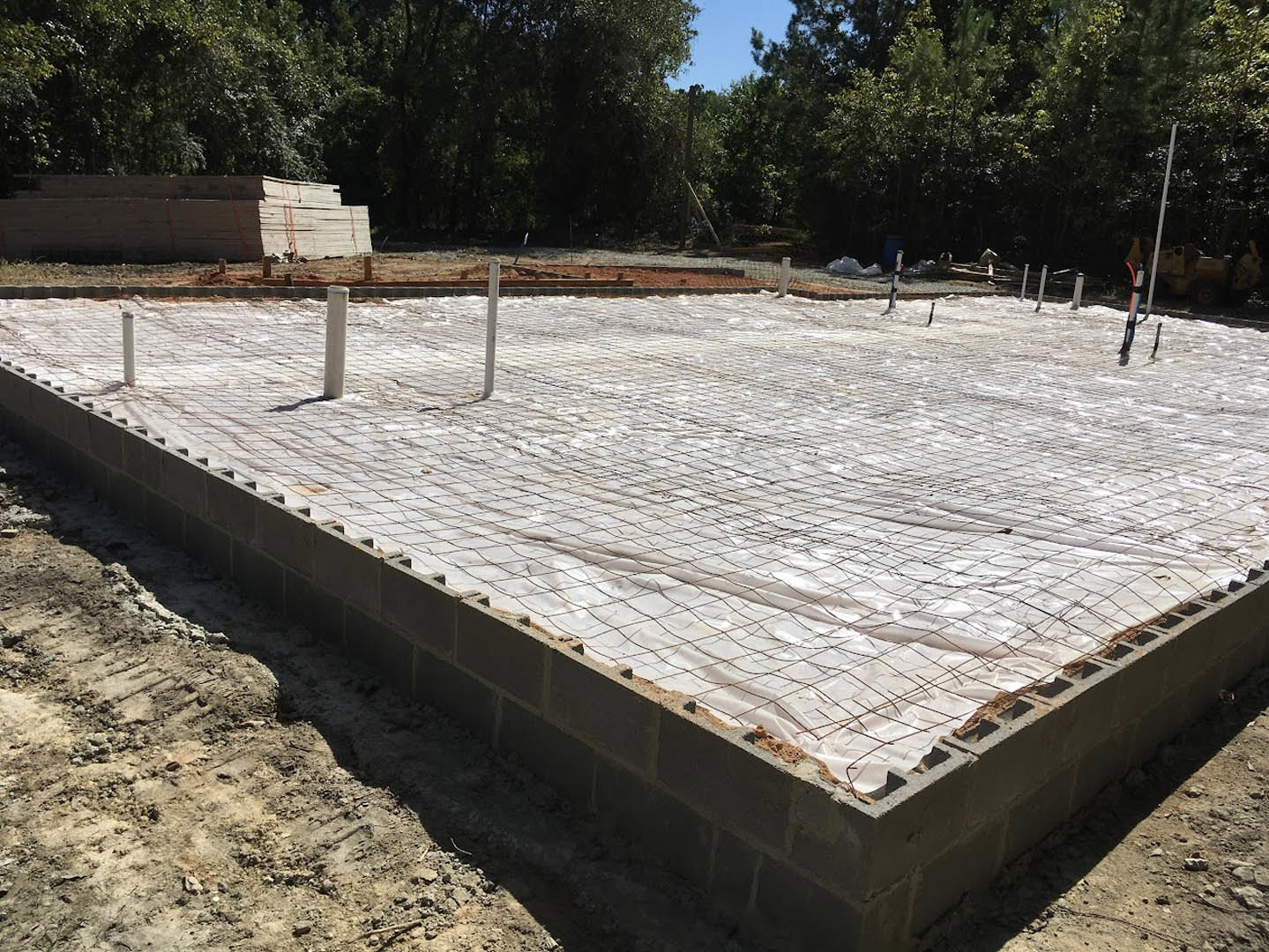 Concrete foundation covered with white plastic sheeting, stack of wood in sunlight, tree with debris pile, white pole with red and blue object, wood stacked in front of trees