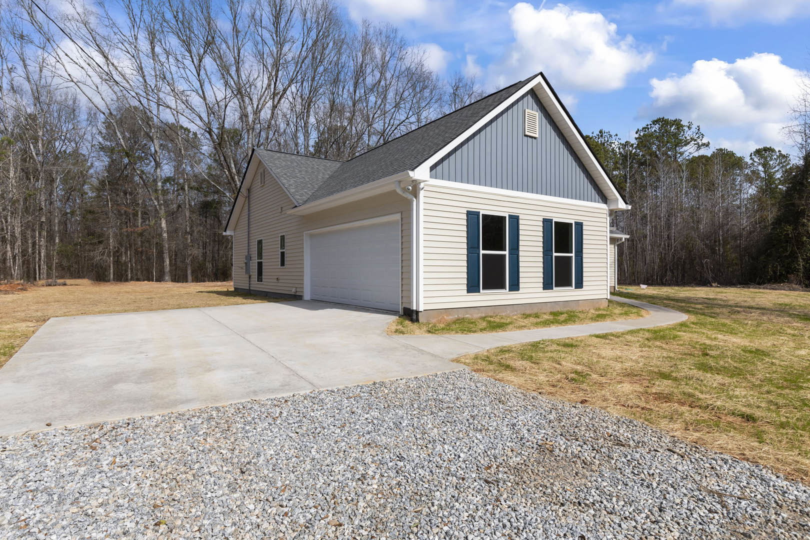 Two-story house with white-framed windows, attached garage featuring paneled door, concrete driveway bordered by gravel, light-colored siding, and blue sky with scattered clouds