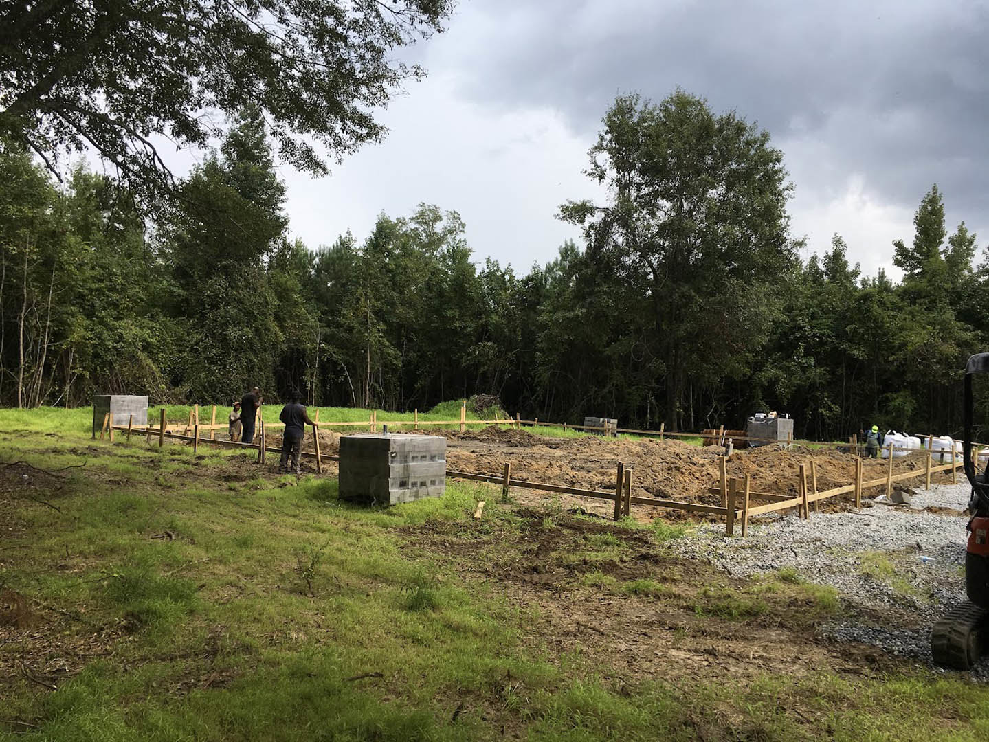Partially built custom home foundation with concrete blocks set in grassy field, several workers present, surrounded by tall leafy trees under cloudy sky