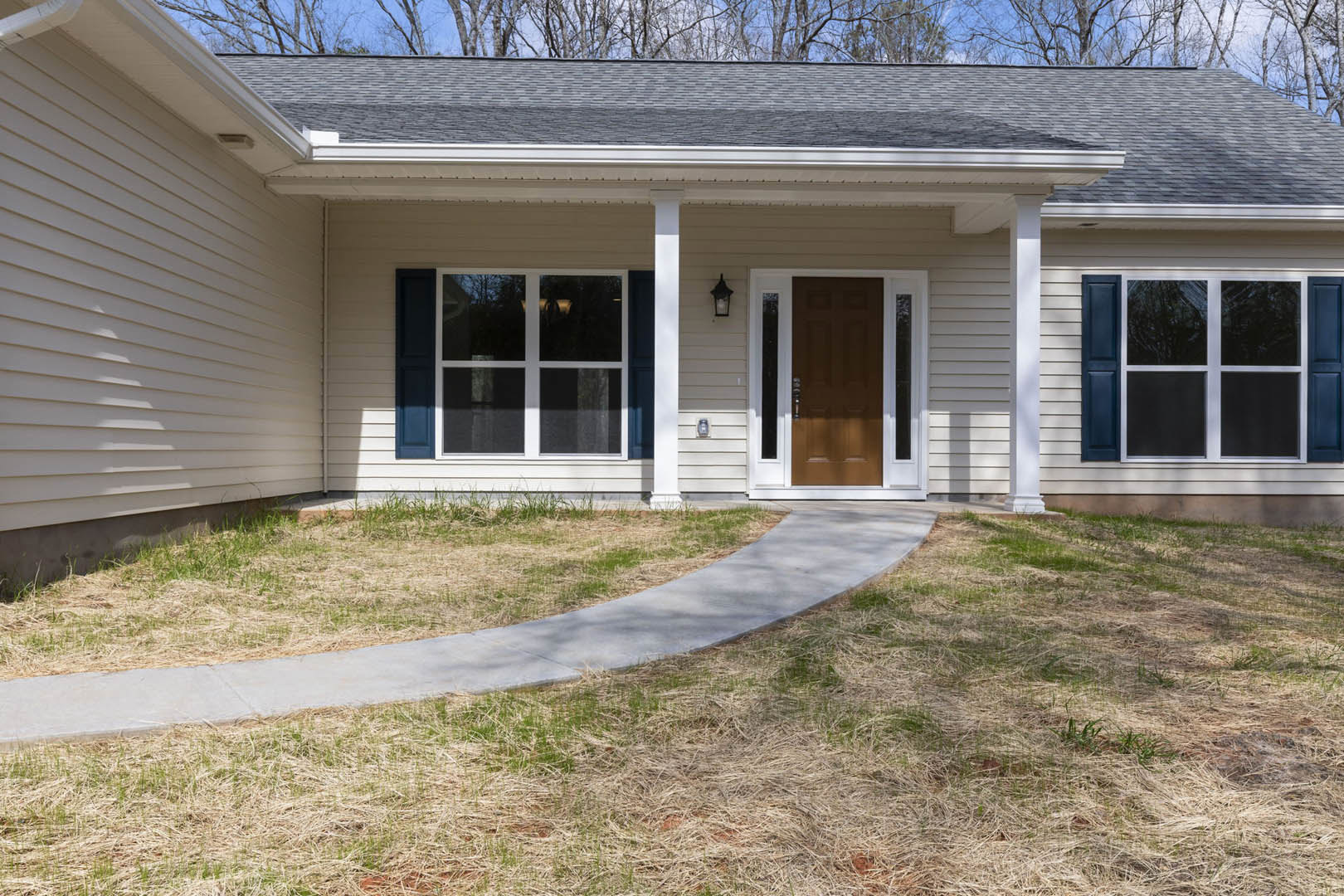 Concrete walkway bordered by green grass leading to a brown front door with glass panels, white siding exterior, and covered porch with windows